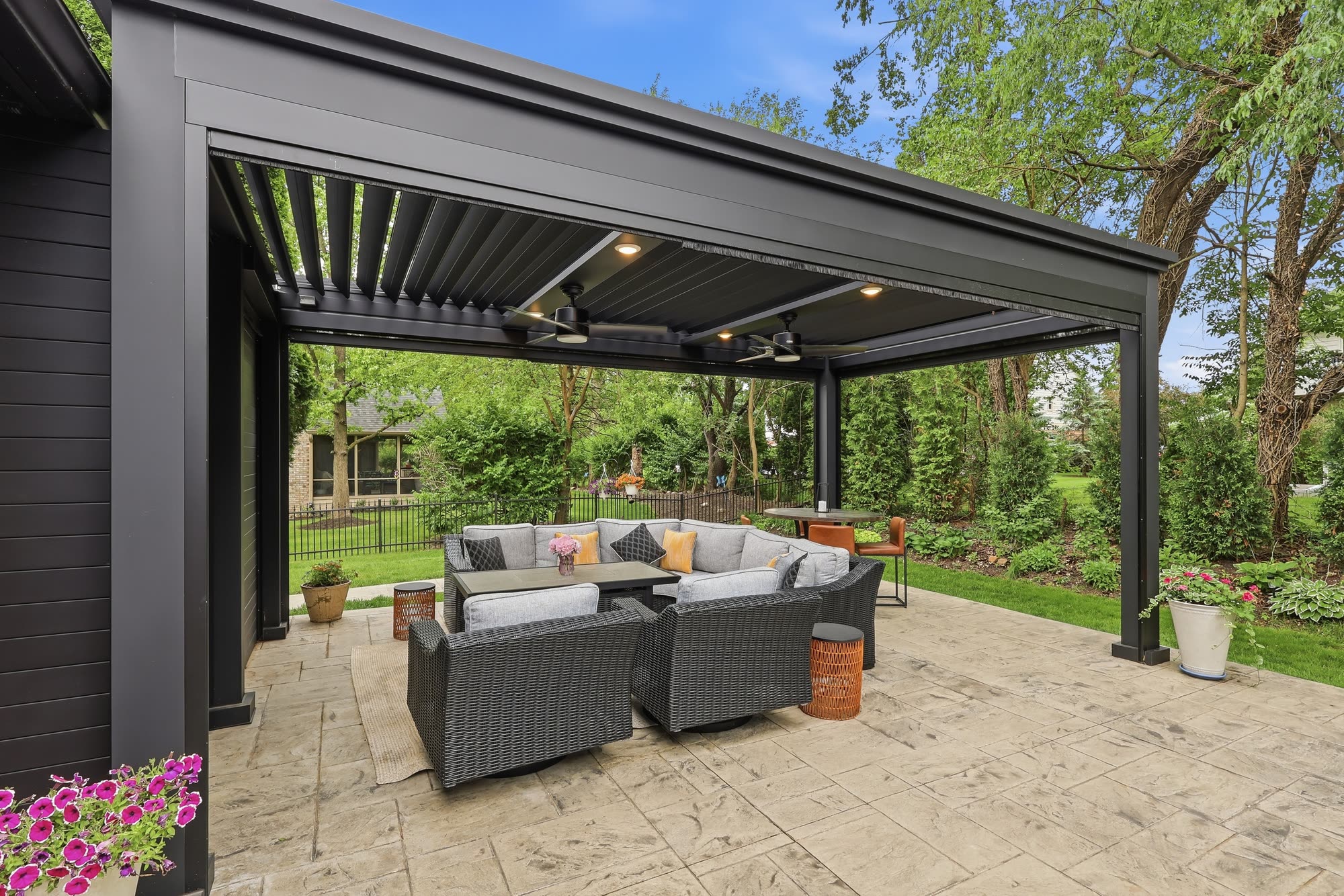 A black louvered pergola with recessed lights and ceiling fans over a stamped concrete patio with an outdoor lounge set.