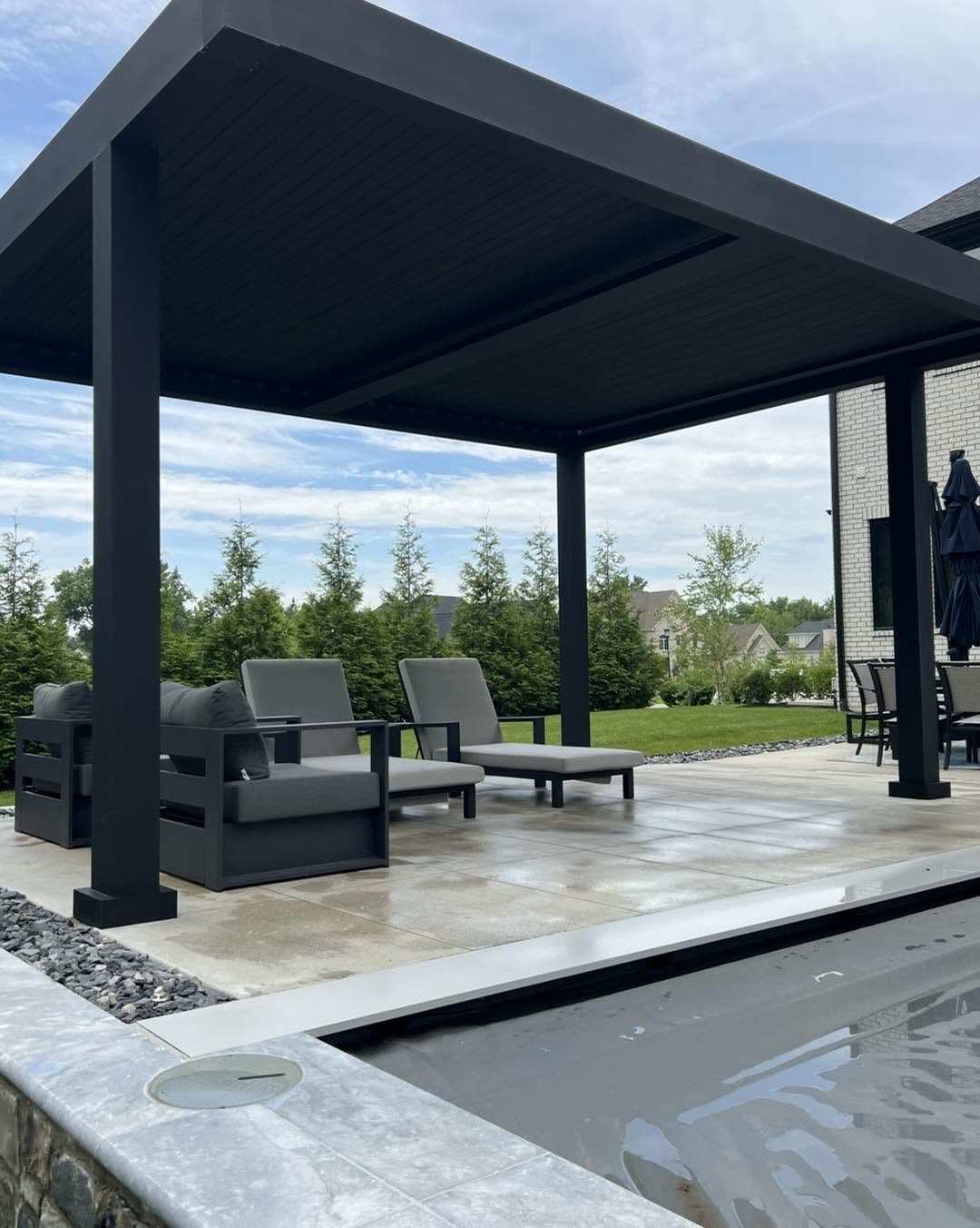 A black louvered pergola shading a lounge seating area on a poolside patio, with a stone-edged pool in the foreground.